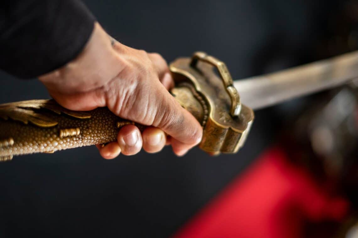 Detailaufnahme einer Hand, die den kunstvoll verzierten Griff eines Katana Schwerts hält – Symbol für japanische Handwerkskunst und traditionelle Schwertkunst.