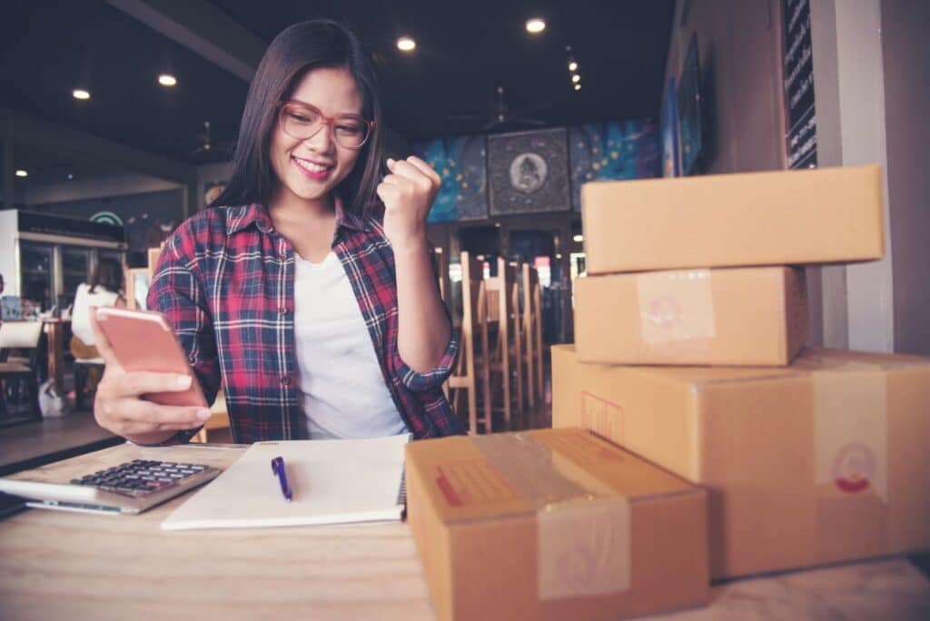 Frau mit Smartphone und Paketen zeigt eine freudige Geste, während sie in einem Café auf ihre Amazon-Bestellungen schaut