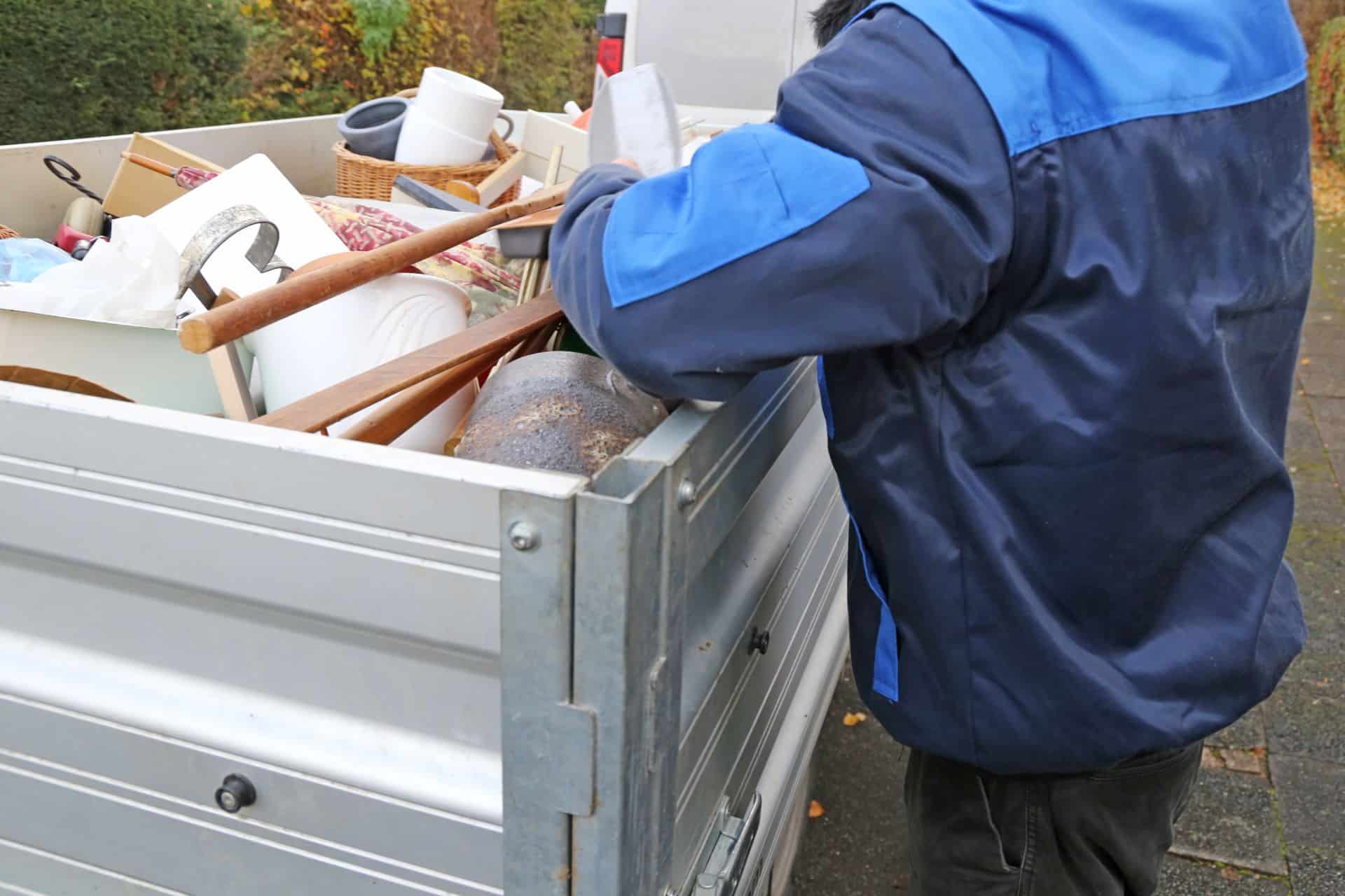 Ein Mitarbeiter in blauer Arbeitskleidung lädt Gegenstände wie Haushaltswaren und Möbelteile auf einen silbernen Transporter für die Entrümpelung.