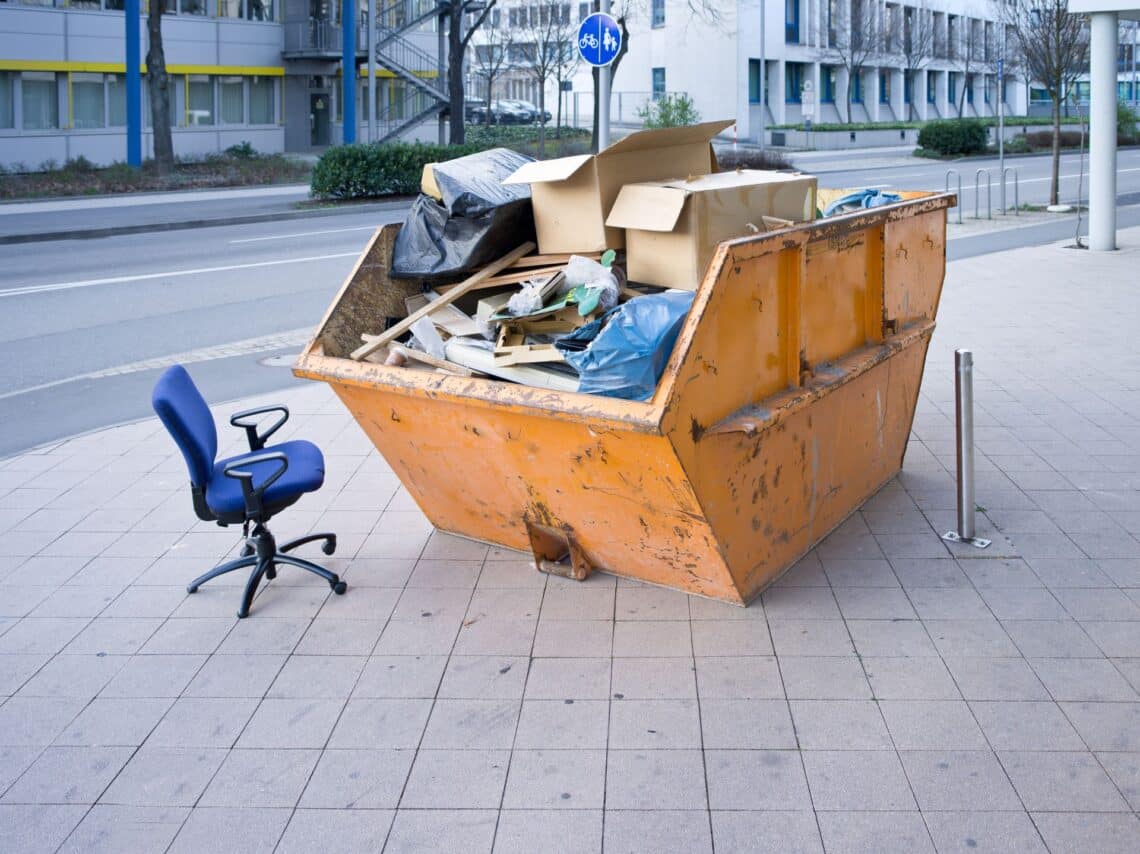 Ein großer orangefarbener Container mit Sperrmüll, darunter Kartons, Holz und Müllsäcke, auf einem Bürgersteig. Daneben steht ein blauer Bürostuhl.