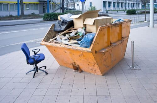 Ein großer orangefarbener Container mit Sperrmüll, darunter Kartons, Holz und Müllsäcke, auf einem Bürgersteig. Daneben steht ein blauer Bürostuhl.