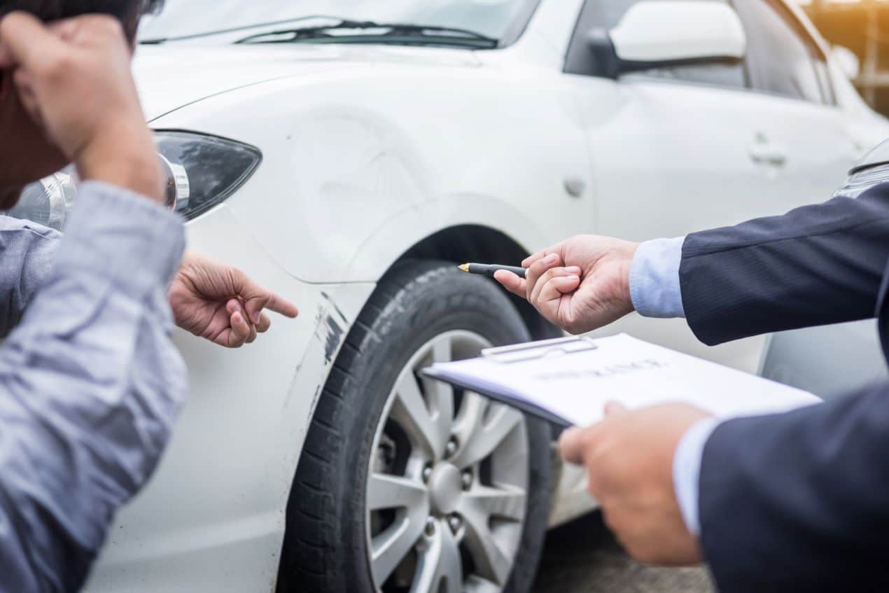 Zwei Personen begutachten einen Unfallschaden an einem Auto, während ein Anwalt mit Klemmbrett und Stift rechtliche Details klärt – Rechtsanwälte Mönchengladbach.