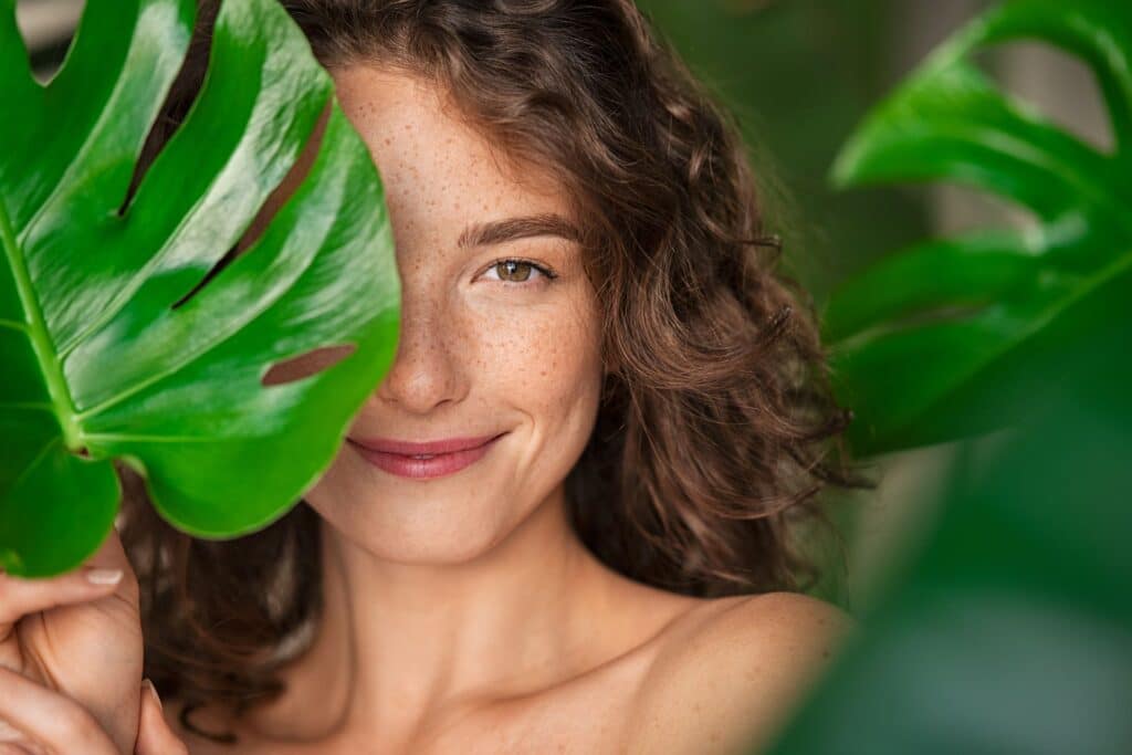 Junge Frau mit ungeschminktem Gesicht blickt durch ein Monstera-Blatt.