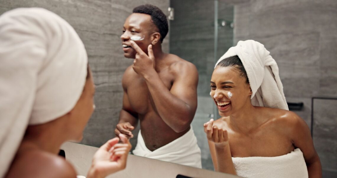 Zwei Personen tragen Gesichtspflege im Badezimmer auf und lachen dabei vor dem Spiegel